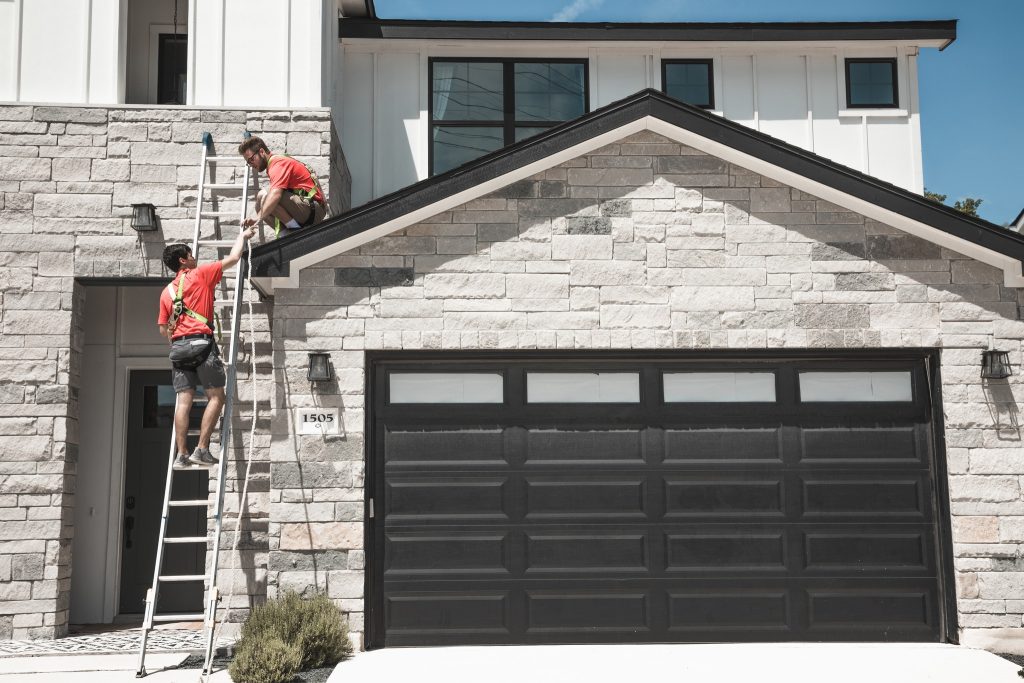 Two workers in safety harnesses install a gutter system on a modern stone house with a dark garage door
