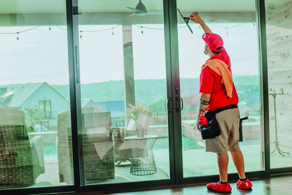 A window cleaner wearing red cleans a large glass door overlooking a patio with a pool. TITLE