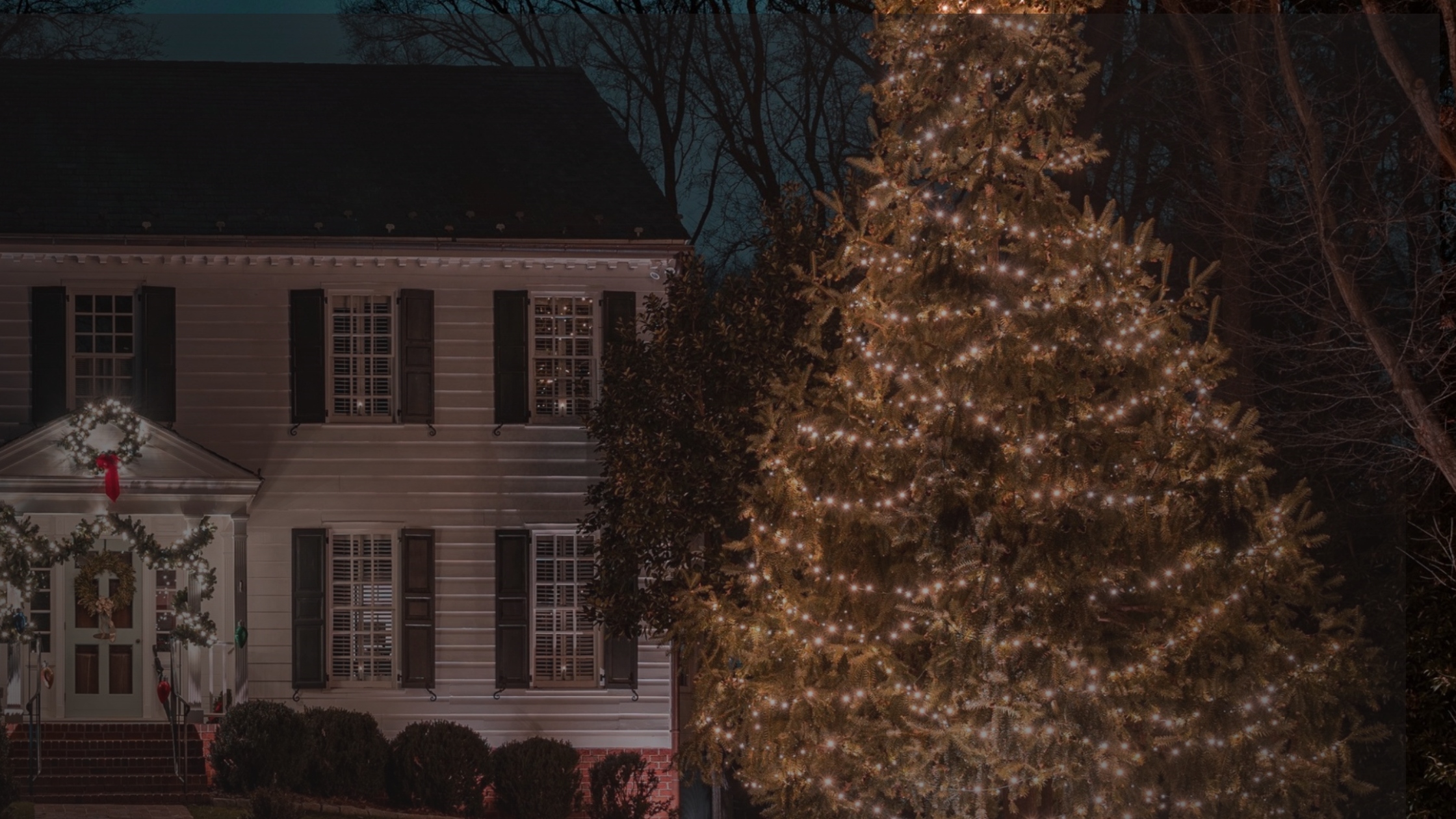 A colonial house decorated with Christmas lights and wreaths stands beside a large, illuminated tree
