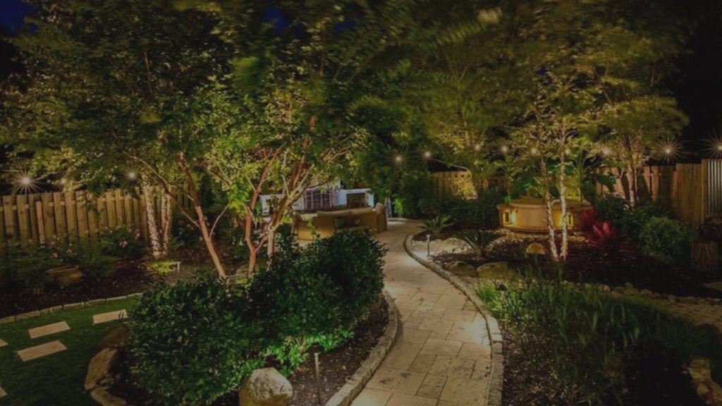 A beautifully illuminated backyard at night with a winding stone path, lush trees, and an outdoor