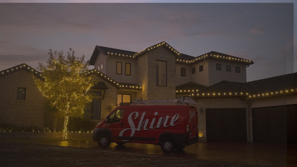 A red Shine van is parked outside a modern home and tree illuminated with warm string lights at