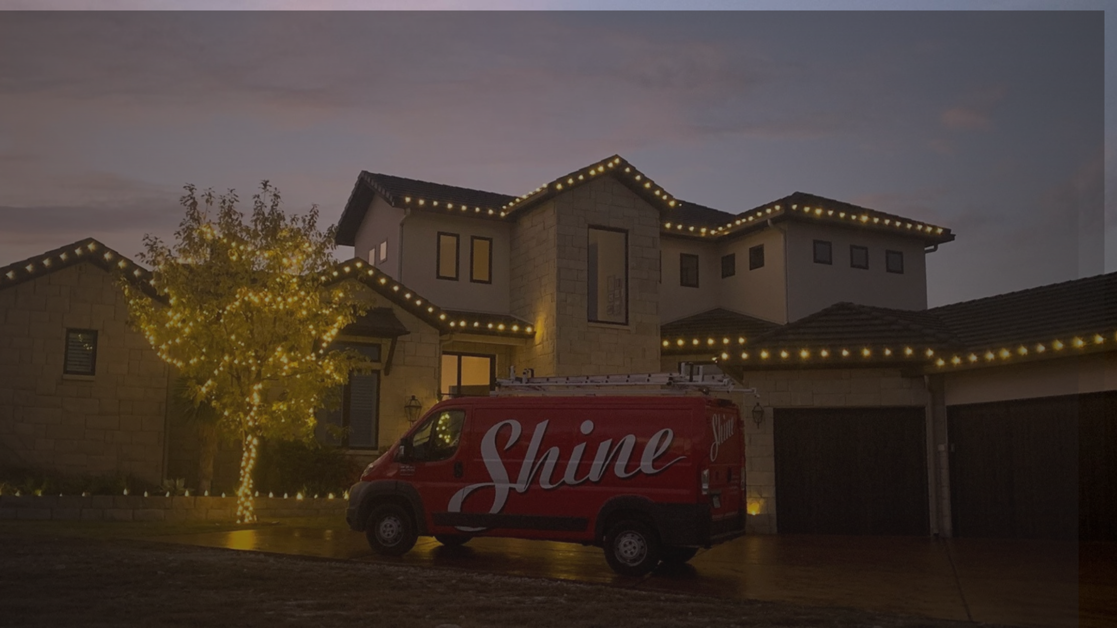 A red Shine van is parked outside a modern home and tree illuminated with warm string lights at