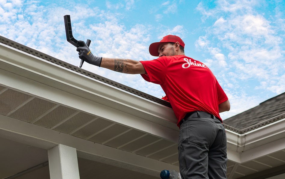 A worker in a red Shine shirt uses a tool to service a house gutter. TITLE: