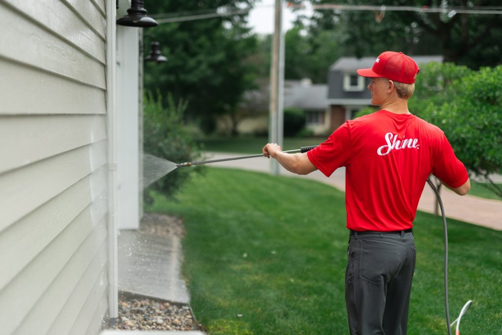 A worker in a red "Shine" shirt power washes the light-colored siding of