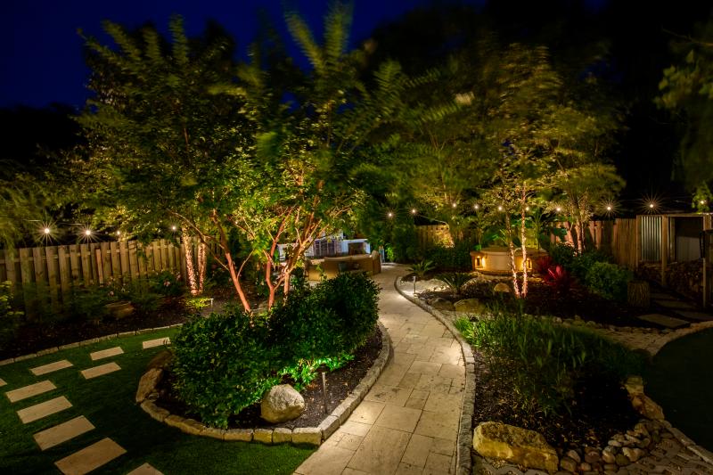 An elegant backyard landscape at night features a winding stone path, glowing trees, and cozy ambient lighting