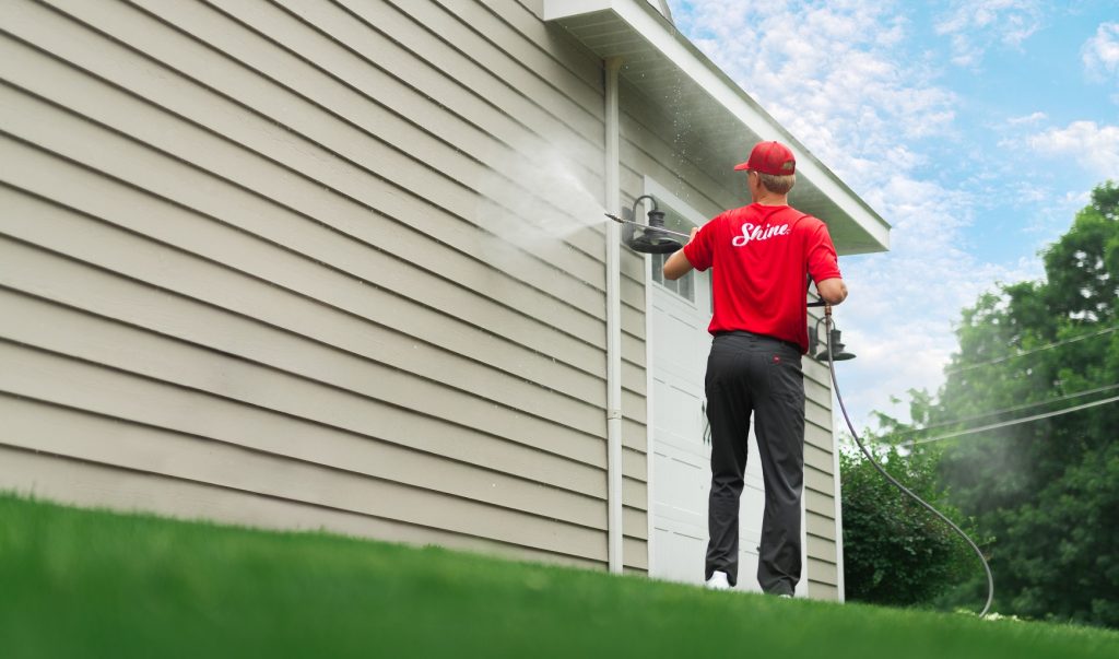 A professional in a red "Shine" shirt power washes the tan siding of a residential