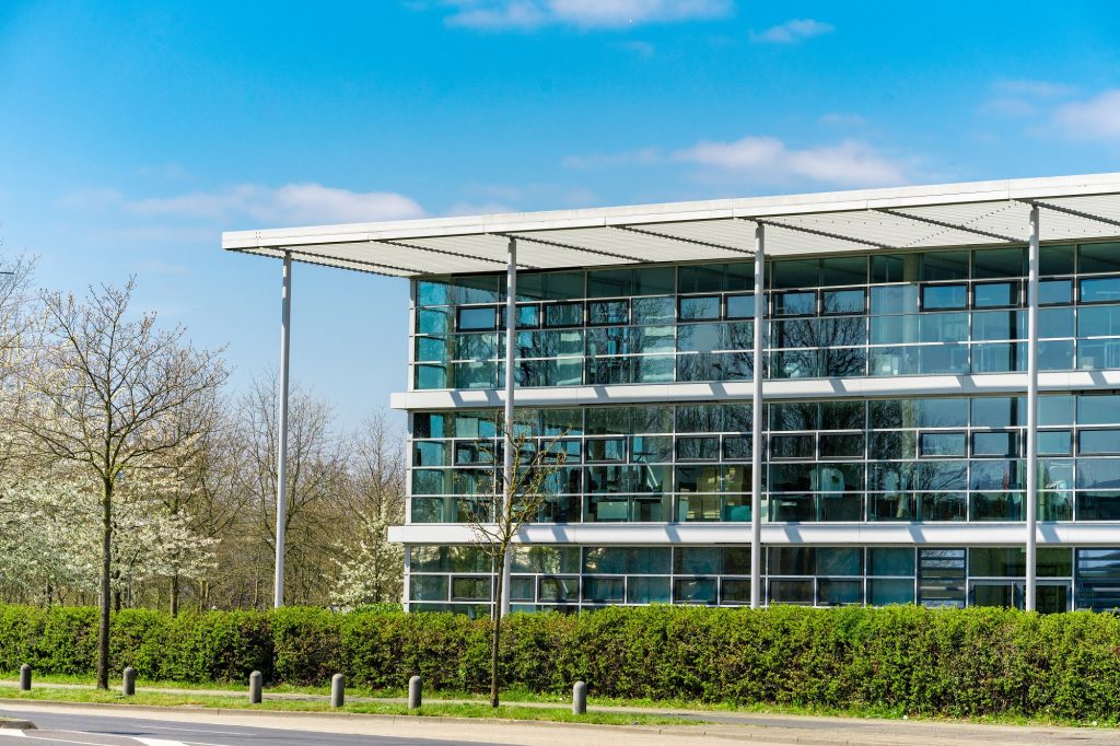Modern multi-story office building with extensive glass windows and green hedges under a blue sky