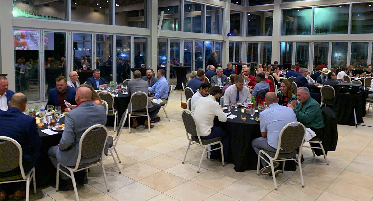 Guests in business attire are seated at round tables enjoying a dinner event in a modern hall.