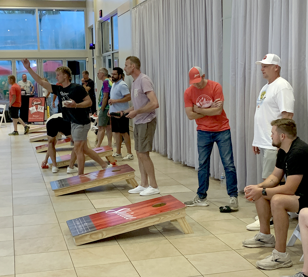 A group of men play cornhole on "Shine" branded boards indoors, with one player