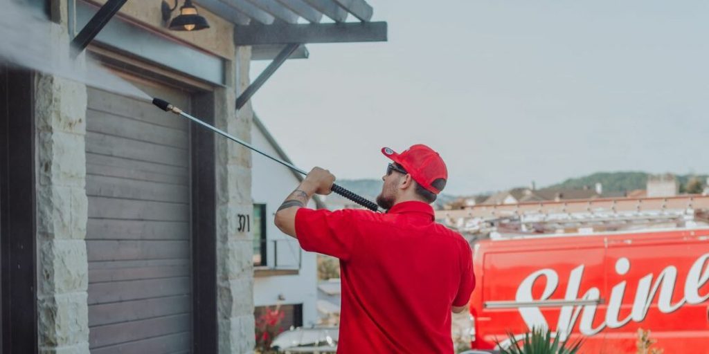A worker in a red uniform uses a power washer to clean a building's stone and wood