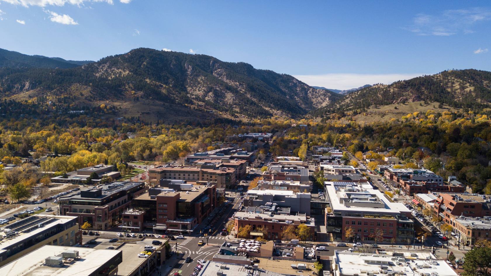 A sunny aerial view of a city nestled against mountains ablaze with golden autumn foliage. TITLE: