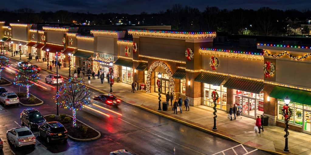A bustling outdoor shopping center glows with holiday lights and decorations at night. TITLE: