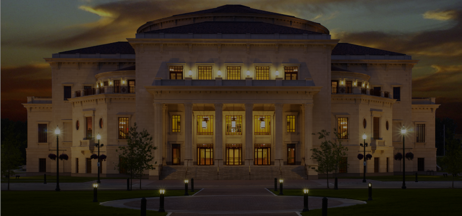 The grand classical building glows with warm light at dusk under a dramatic sky. TITLE: