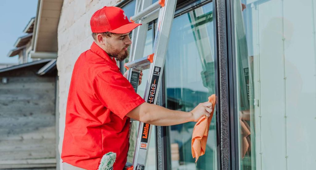 A man in a red uniform cleans a large glass window from a ladder.
