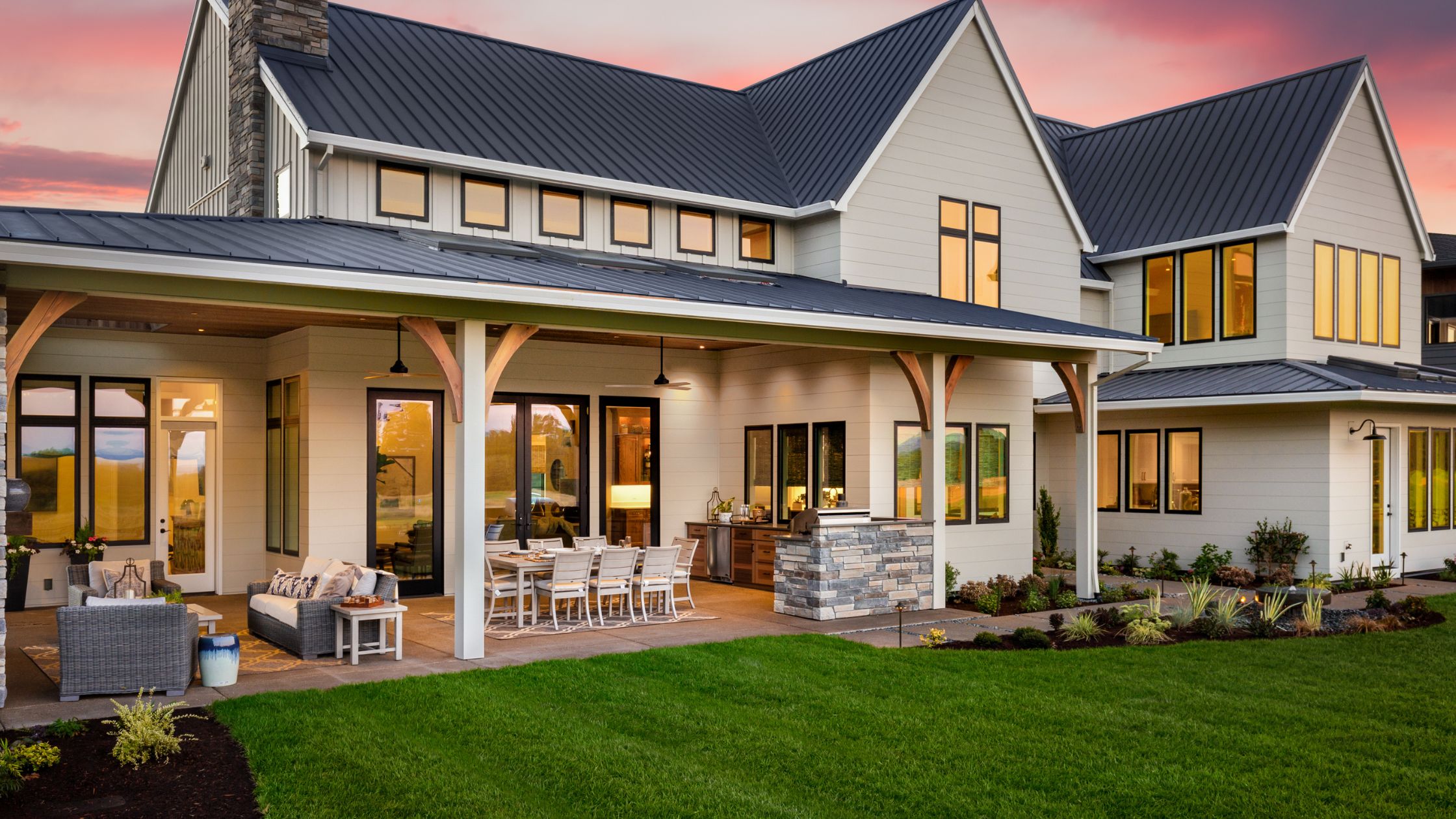 Elegant modern farmhouse with a covered patio, outdoor kitchen, and lush lawn at sunset.