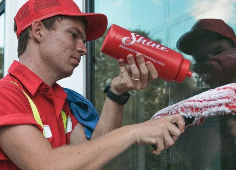 A window cleaner in a red uniform applies solution from a Shine bottle to a large window.