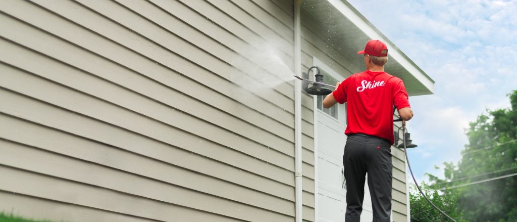 A professional in a red uniform power washes the beige siding of a house.