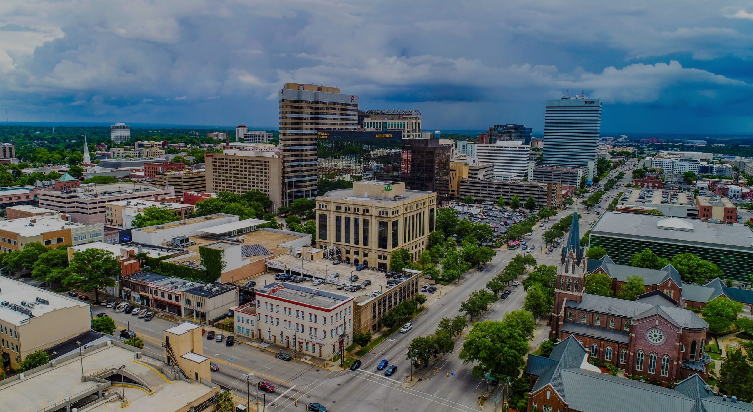 Aerial view of downtown Columbia, South Carolina, showing skyscrapers, streets, and churches under a cloudy