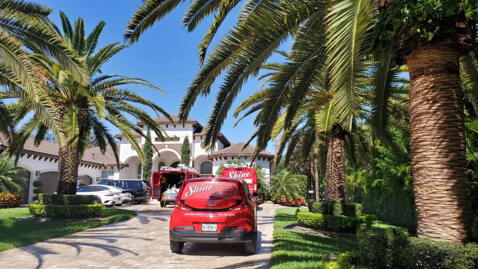 Two red Shine service vehicles are parked on a paved driveway in front of a large, palm