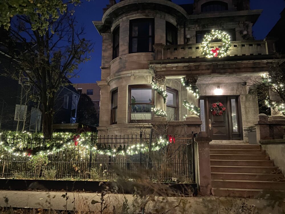 A grand stone house glows with festive Christmas lights and wreaths against a dark evening sky.