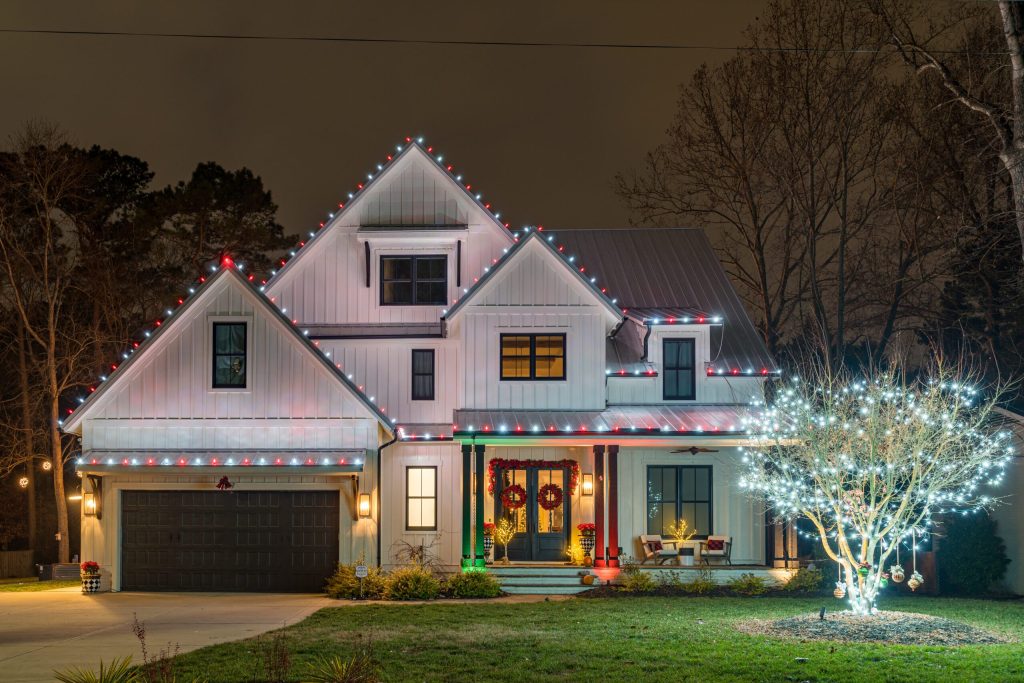 A festive modern farmhouse glows at night with red and white Christmas lights and a sparkling tree.