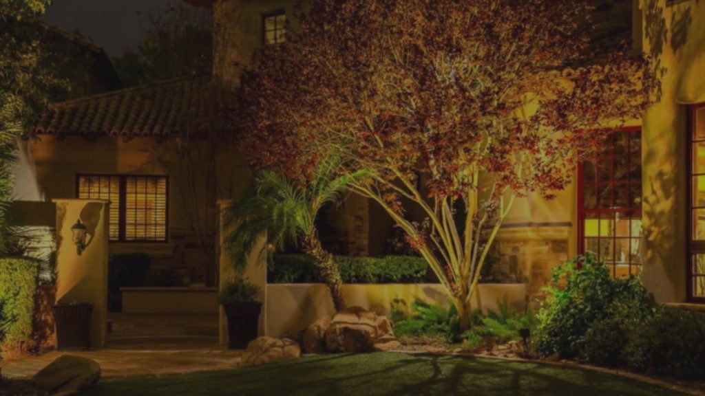 Warm landscape lighting illuminates a Mediterranean-style house and a vibrant red-leafed tree at