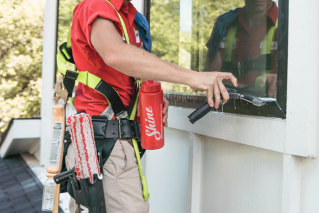 A professional window cleaner in a red uniform and safety harness uses a squeegee on a window.