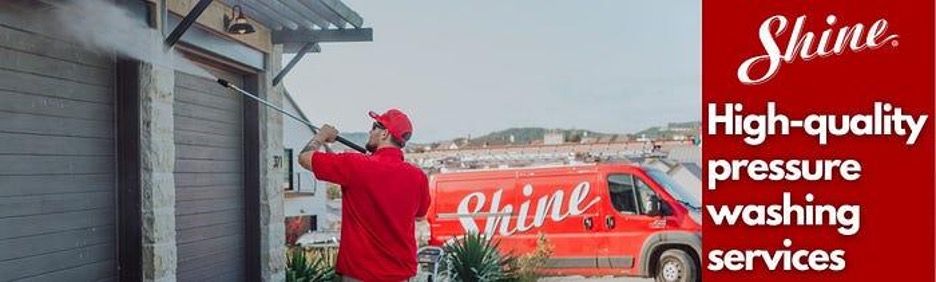 A worker in a red uniform pressure washes a building wall next to a Shine service van.