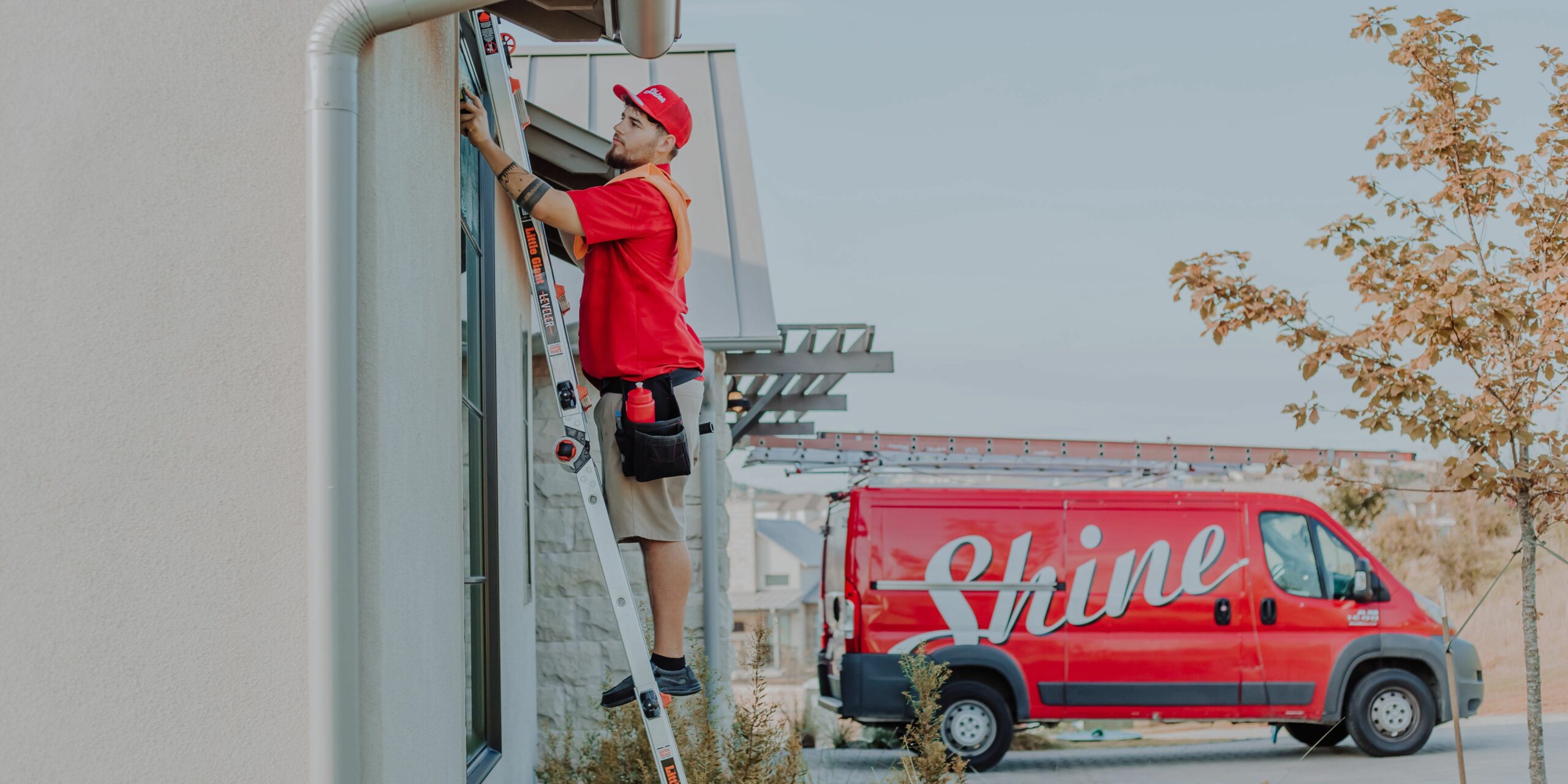 A worker on a ladder cleans a building's window with a red Shine service van parked nearby