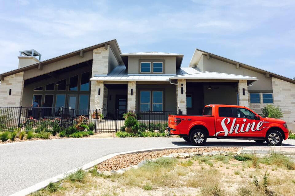 A bright orange Shine pickup truck is parked on the driveway in front of a modern, stone and stucco house.