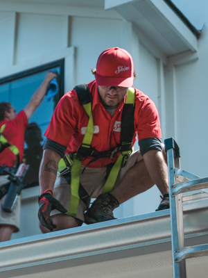 A worker in a red uniform and safety harness squats on a roof, with another cleaning