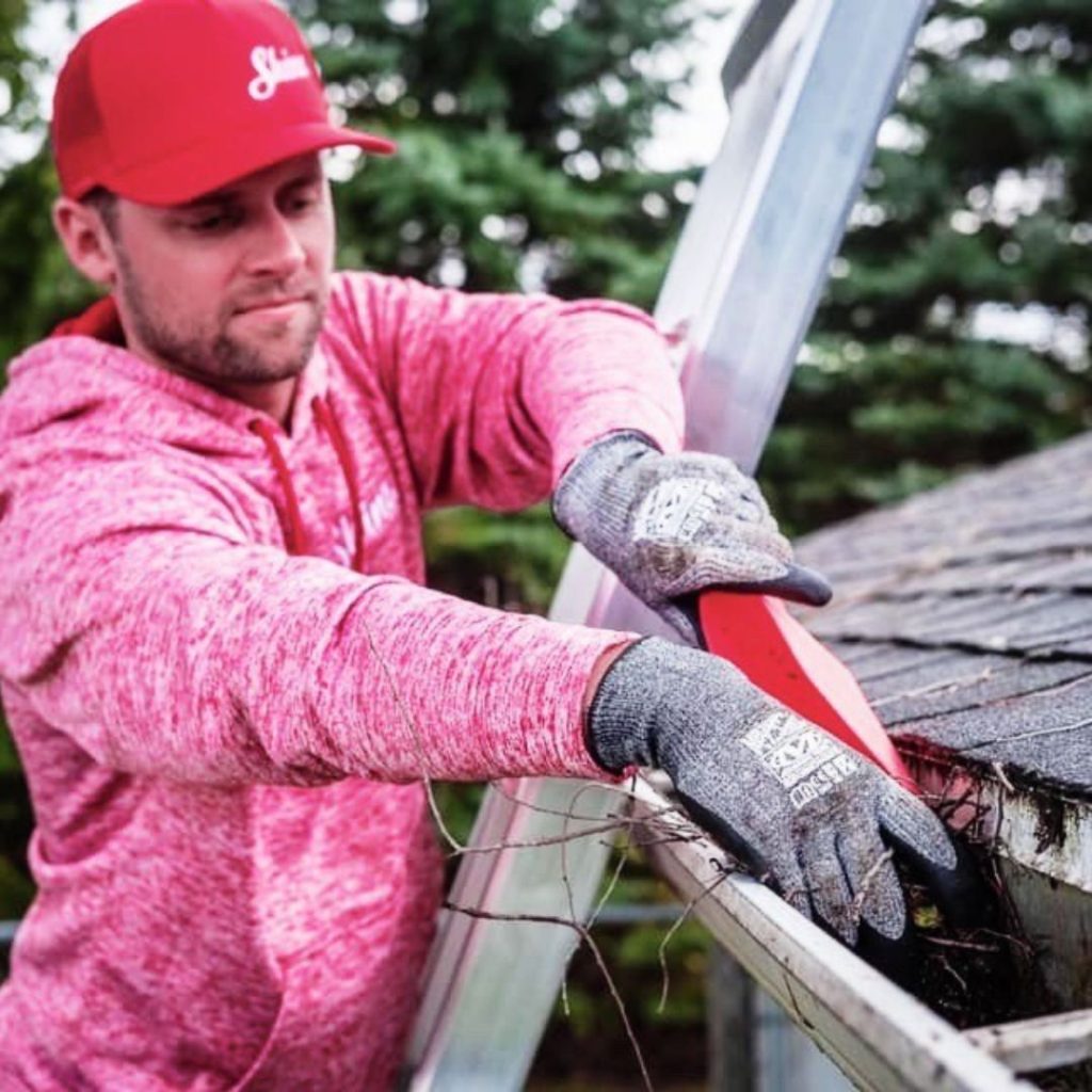 A man in a red hat and gloves stands on a ladder, cleaning a clogged