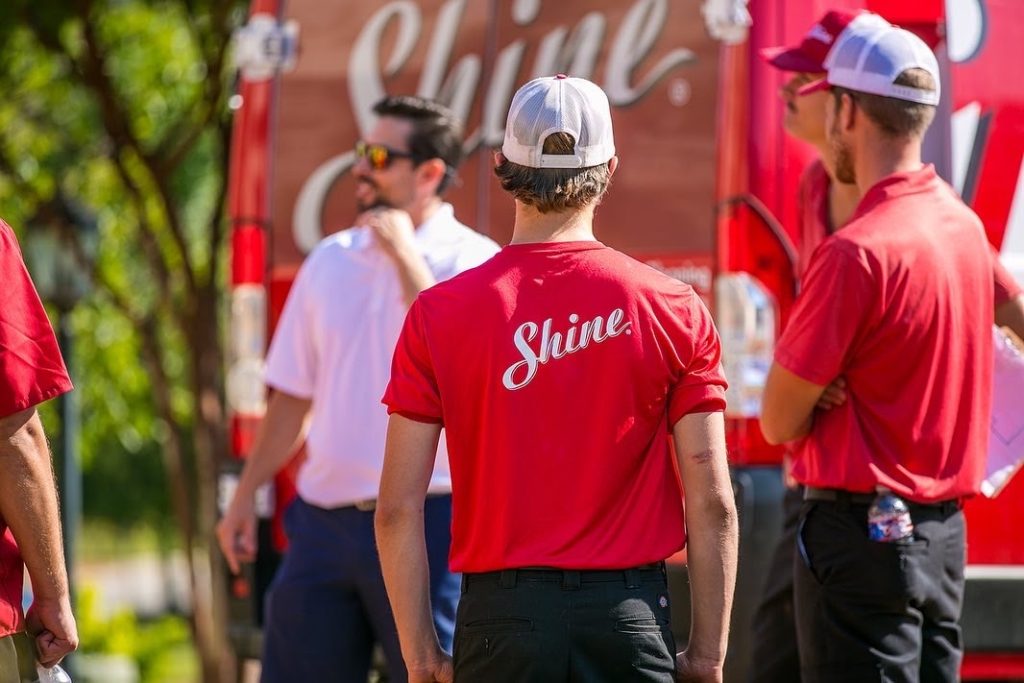 A person in a red "Shine" shirt and white cap stands with a