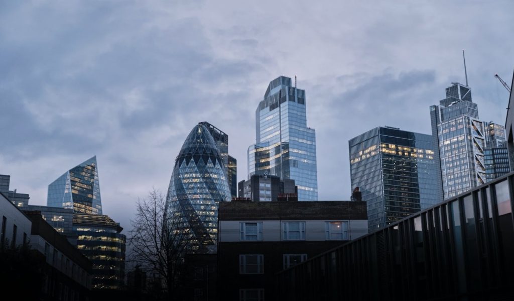 Modern London skyscrapers, including the Gherkin, stand tall against a cloudy sky with illuminated
