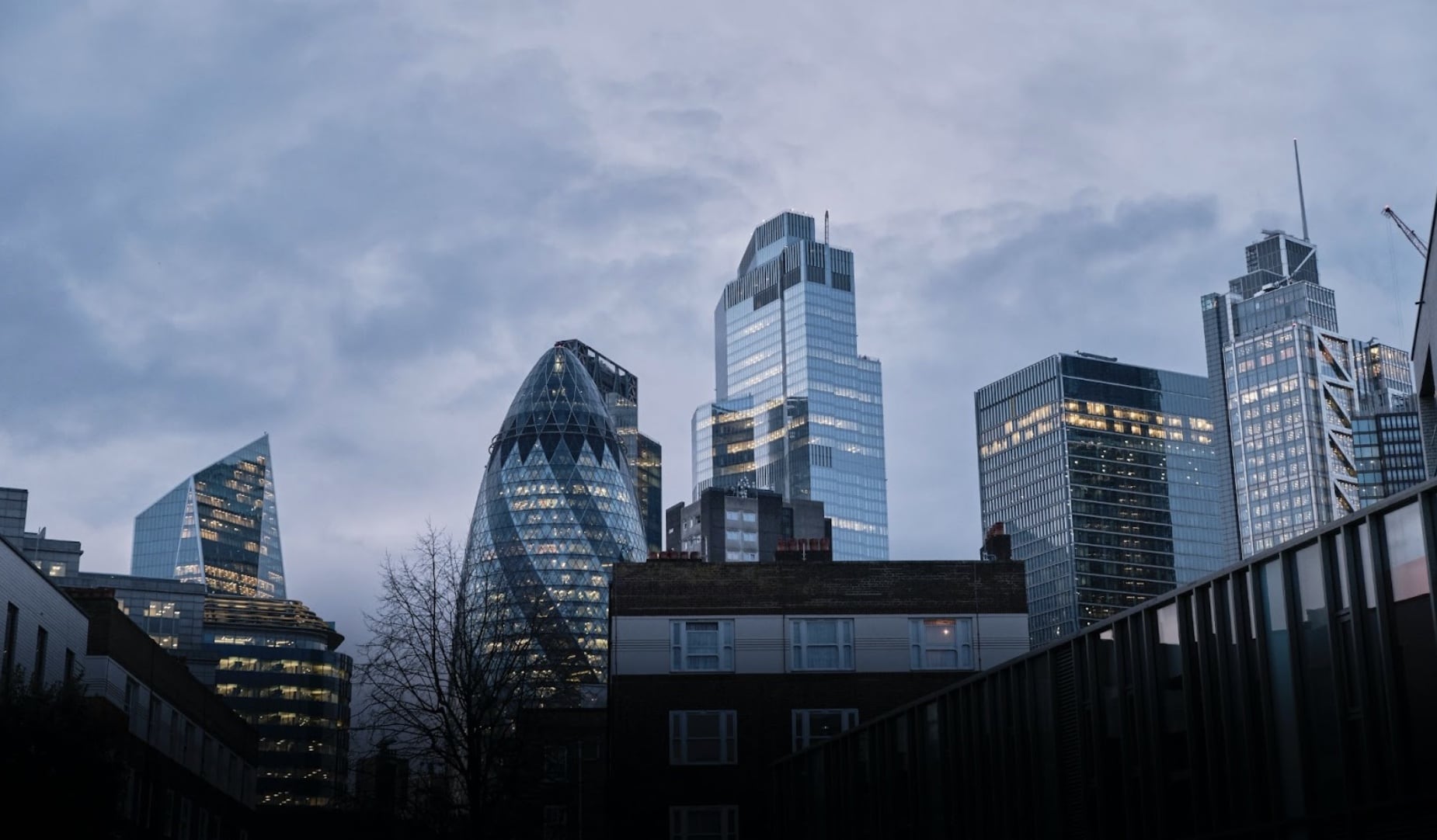 Modern London skyscrapers, including the Gherkin, stand tall against a cloudy sky with illuminated