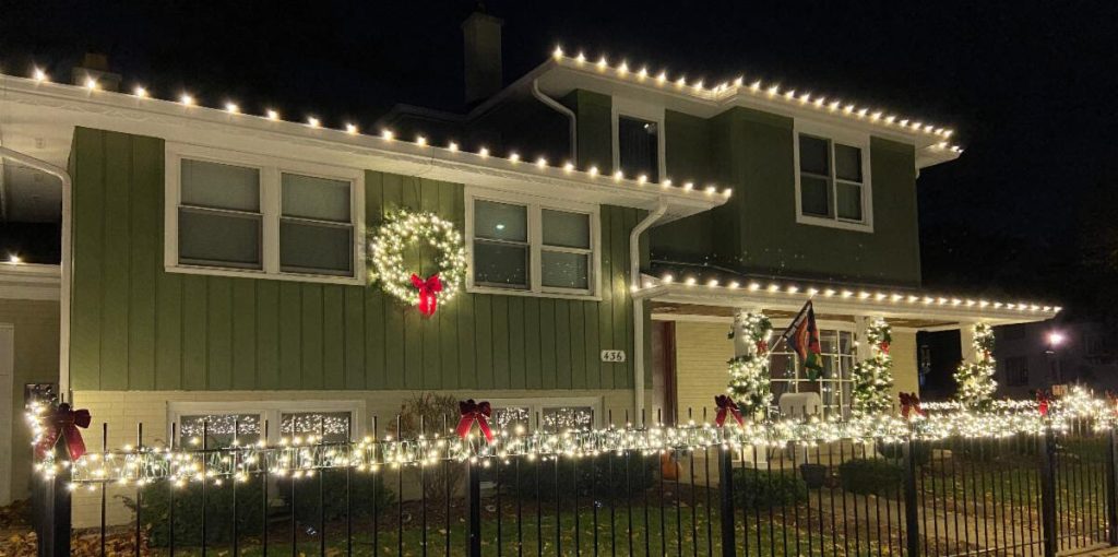 A green house is brightly decorated with white Christmas lights and a large lit wreath at night.