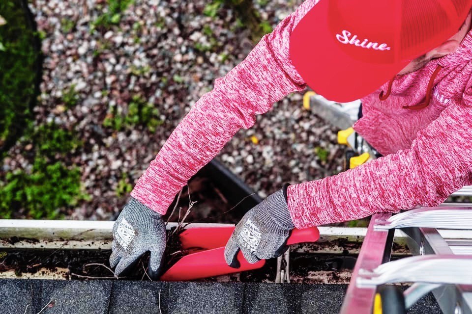 A person wearing a red hat and gloves cleans a dirty roof gutter with a red scoop