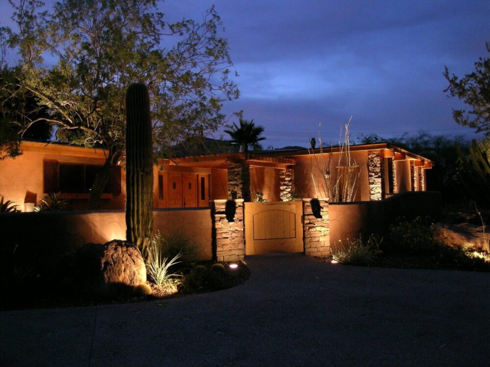 A desert-style home glows warmly at dusk, illuminated by landscape lighting highlighting a saguaro cactus and stone walls.