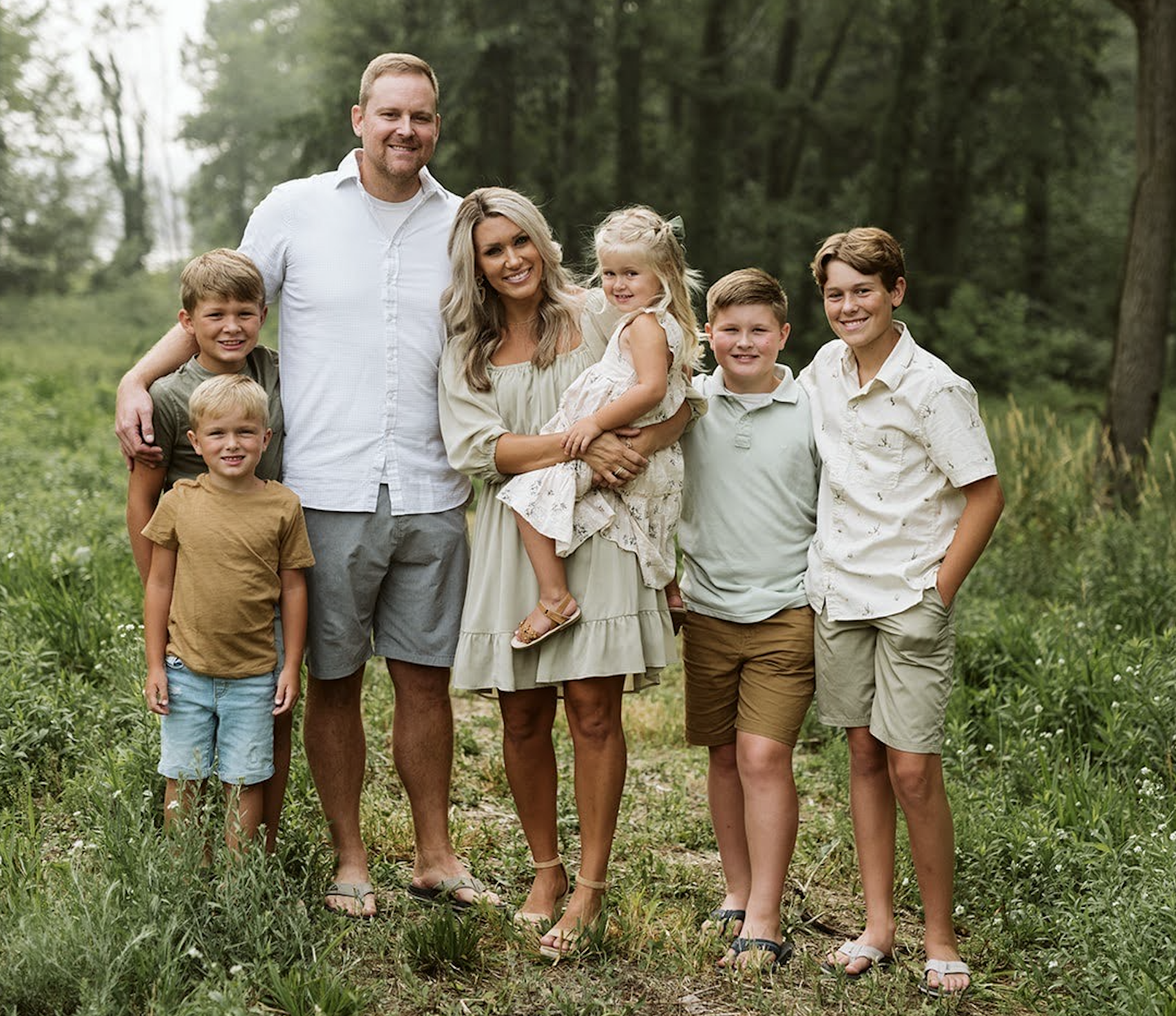 A smiling family of six, including parents and four children, stands in a grassy outdoor setting.