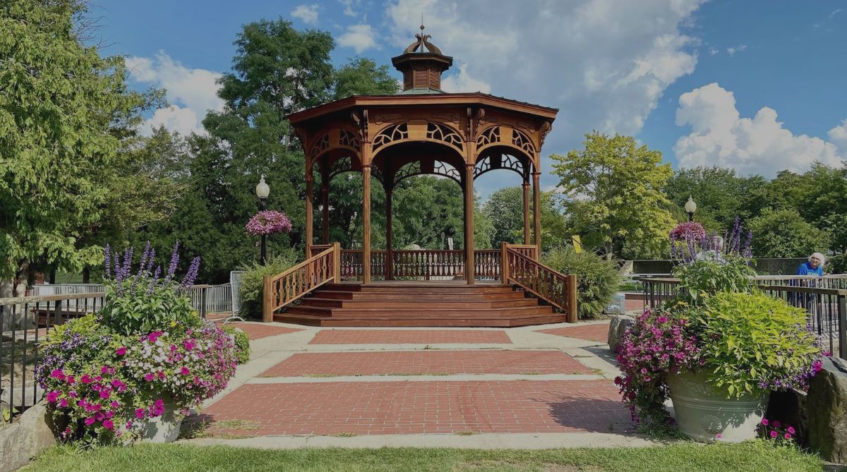 Ornate wooden gazebo stands in a vibrant park with blooming flowers under a bright blue sky.