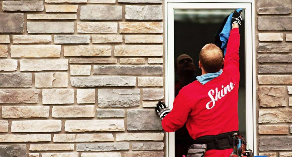 A person in a red "Shine" shirt cleans a window on a stone building.