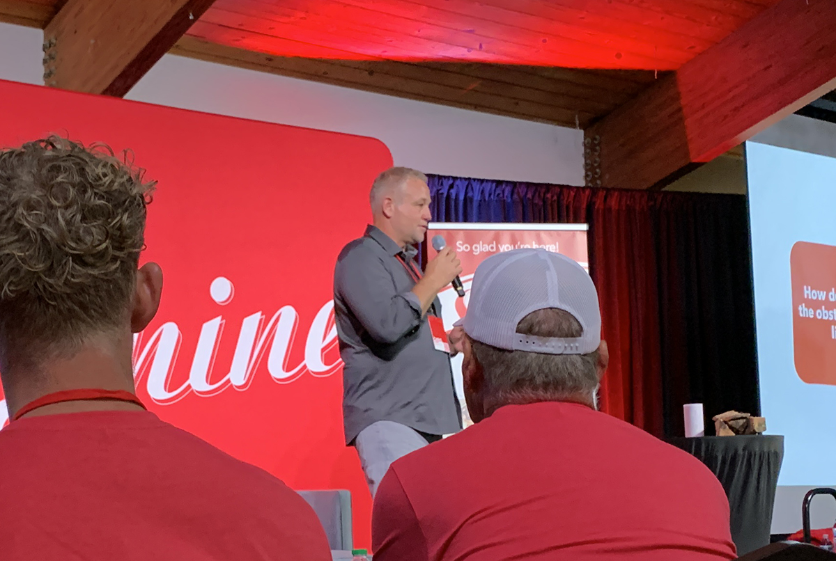 A man speaks on a stage with a red backdrop and screen, viewed from the