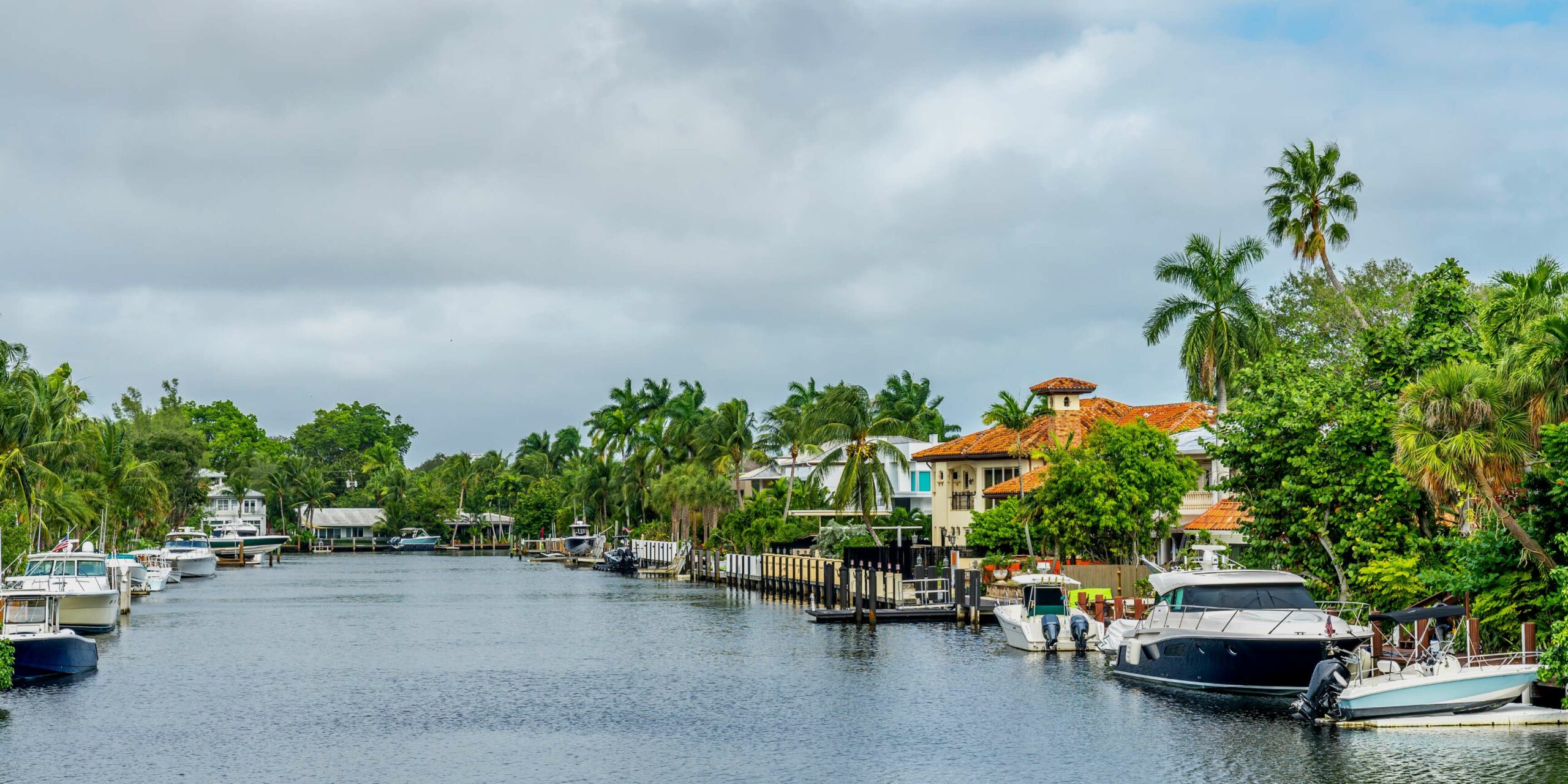 Luxury boats are moored along a tropical canal lined with waterfront homes and lush palm trees.