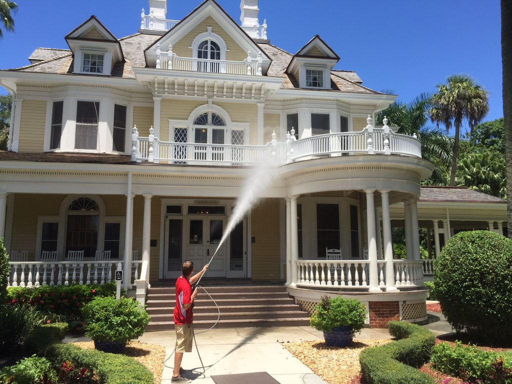 A person power washes the ornate yellow facade of a large Victorian house on a sunny day.