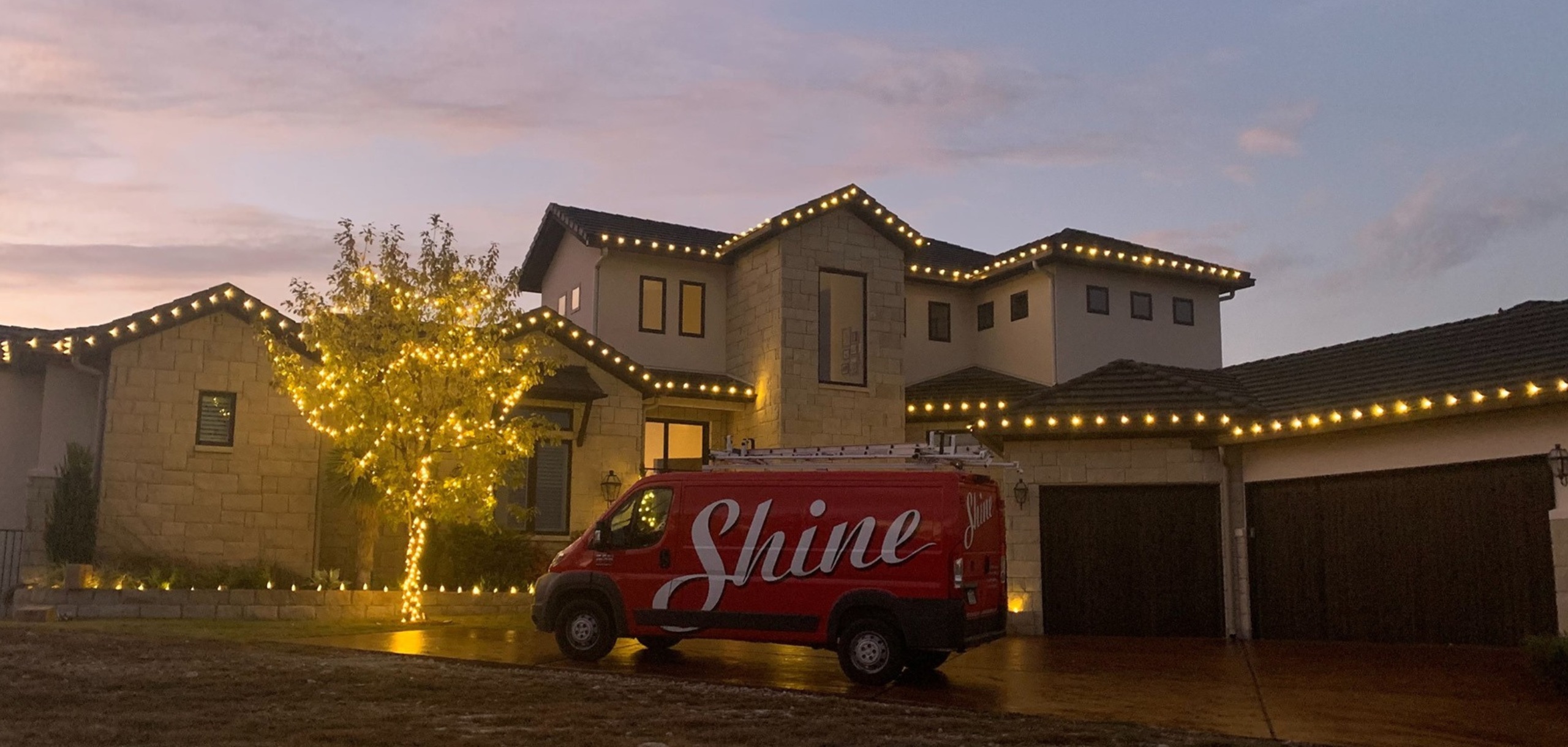 A red Shine van is parked in front of a large house beautifully decorated with warm white string
