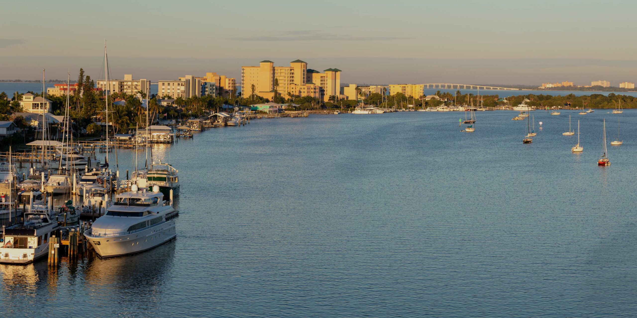 A picturesque coastal town features a bustling marina, numerous boats, and a bridge spanning the tranquil bay