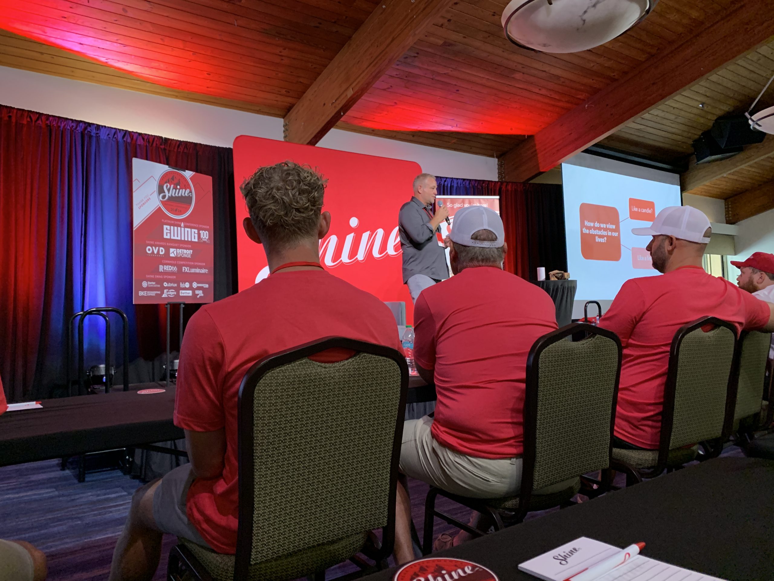 An audience watches a speaker on stage at a conference with a 'Shine' banner