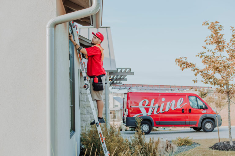 A worker on a ladder cleans a window next to a red "Shine" service van.