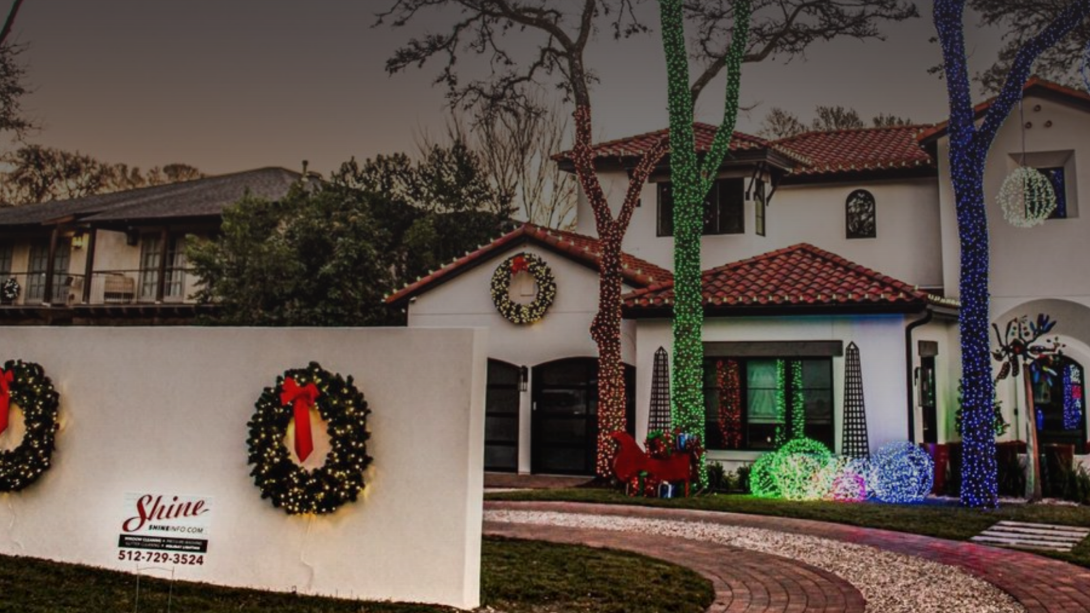 A large house is festively decorated with colorful Christmas lights, wreaths, and yard displays