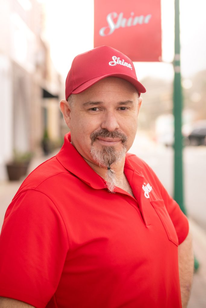 Smiling man in a red "Shine" polo shirt and cap with a tied goatee stands outdoors